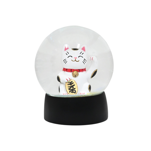 A clear glass snow globe featuring a white Maneki-neko (lucky cat) figurine with pink ears, a red collar, and a gold bell, sitting on a black cylindrical base against a white background.