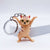 A small cat-shaped keychain charm standing on its hind legs with arms raised, attached to a silver metal key ring. The cat is light peach-beige with pink and dark brown stripes, yellow eyes, and a black nose, photographed against a clean white studio background.