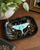 A decorative black tray with a gold-toned fairy design and a large light blue luna moth, styled on a wooden table with jewelry and a plant.