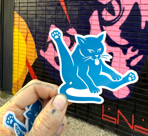 Blue cat-shaped sticker held by a hand in front of a colorful graffiti wall.