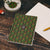 Notebook with cannabis leaf pattern and cover reads "I feel amazingly creative today." Notebook sits on a wooden desk with a marijuana cigarette nearby. 