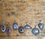 Six handmade fordite pendant necklaces with silver chains are arranged on a rustic wooden surface. The pendants feature various abstract concentric swirl patterns in black, white, red, and blue.