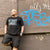 Man wearing a black 'Detroit Famous' t-shirt leaning against a wall with graffiti. Jeferson Ciarvi.