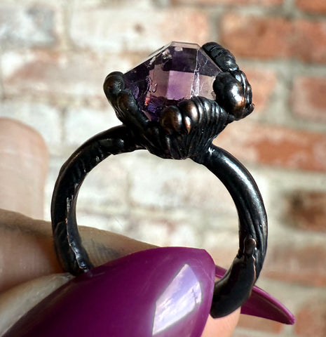 A close-up of a gothic-style ring with a large faceted purple gemstone held by dark, claw-like metal fingers, shown being held by a hand with purple nail polish against a blurred brick wall background.