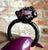 A close-up of a gothic-style ring with a large faceted purple gemstone held by dark, claw-like metal fingers, shown being held by a hand with purple nail polish against a blurred brick wall background.